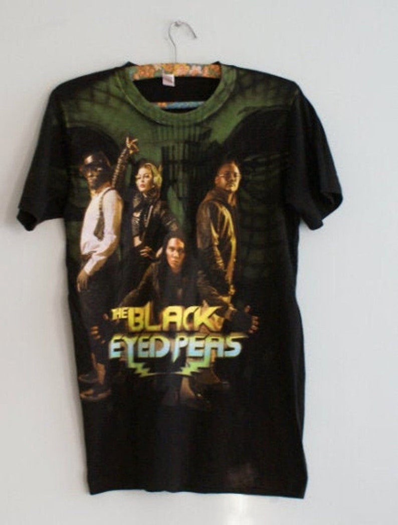 May include: Black t-shirt featuring a graphic of The Black Eyed Peas band members. The design includes the band's name in a stylized font with a green and yellow color scheme. The shirt has a green and black tie-dye pattern on the top.