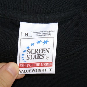 May include: Close-up of a black t-shirt collar with a white tag. The tag features the text "SCREEN STARS by FRUIT OF THE LOOM" and "VALUE WEIGHT T". The tag also includes the size "M" and the text "100% COTTON".