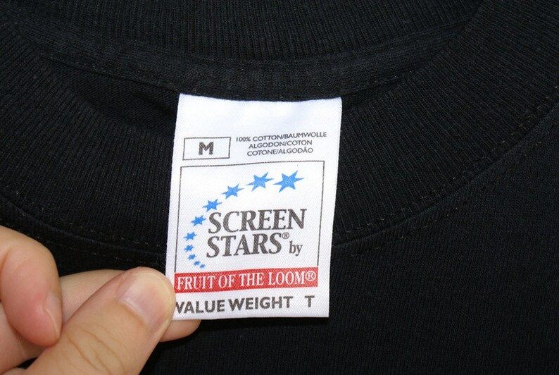 May include: Close-up of a black t-shirt collar with a white tag. The tag features the text "SCREEN STARS by FRUIT OF THE LOOM" and "VALUE WEIGHT T". The tag also includes the size "M" and the text "100% COTTON".