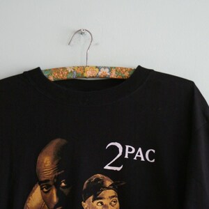 K&ouml;nnte beinhalten: Schwarzes T-Shirt mit einem Grafikdruck von zwei M&auml;nnern. Der Text "2PAC" ist in Wei&szlig; auf dem Shirt gedruckt.