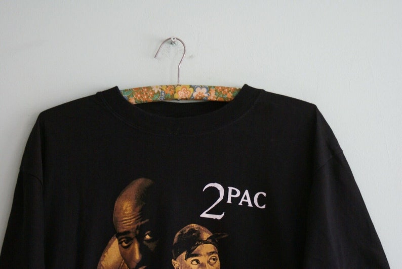 K&ouml;nnte beinhalten: Schwarzes T-Shirt mit einem Grafikdruck von zwei M&auml;nnern. Der Text "2PAC" ist in Wei&szlig; auf dem Shirt gedruckt.