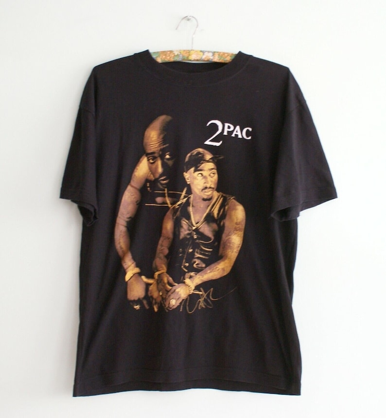 K&ouml;nnte beinhalten: Schwarzes T-Shirt mit einem goldenen Aufdruck von 2Pac. Der Druck zeigt zwei Bilder von 2Pac, eines mit einem schwarzen Bandana und eines ohne. Der Text "2PAC" ist in Wei&szlig; &uuml;ber den Bildern gedruckt.