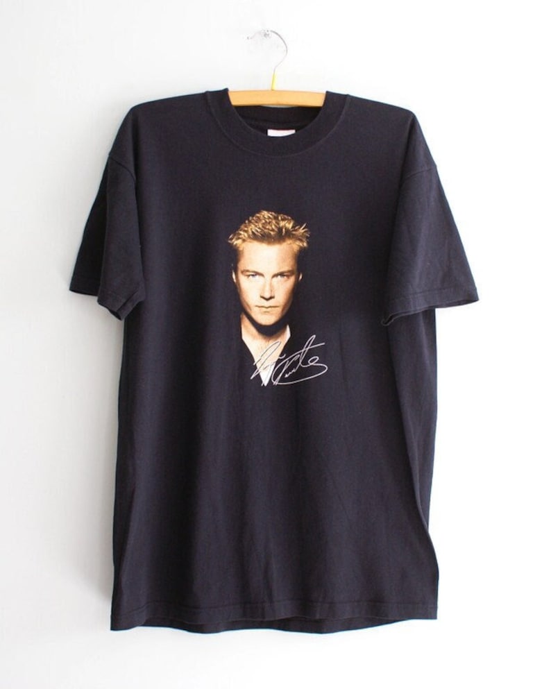 May include: Black t-shirt featuring a portrait of a man with blonde hair and a signature in white. The t-shirt is hanging on a wooden hanger against a white background. The image on the shirt is in shades of brown and gold.