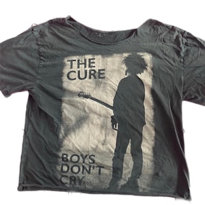 May include: Dark gray t-shirt featuring a graphic of a person with a guitar and the text "THE CURE" above and "BOYS DON'T CRY" below. The shirt has a distressed look with raw edges.