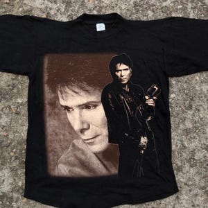 May include: Black t-shirt with a printed image of a man with long dark hair holding a microphone. The image is in a brown frame.