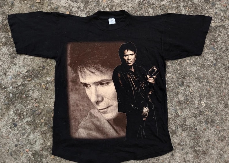 May include: Black t-shirt with a printed image of a man with long dark hair holding a microphone. The image is in a brown frame.