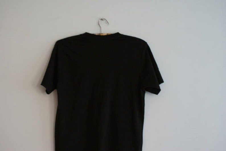 May include: A solid black t-shirt hanging on a metal hanger against a white wall. The short-sleeved shirt is made of a soft material and is a basic wardrobe staple.