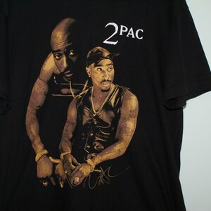 K&ouml;nnte beinhalten: Schwarzes T-Shirt mit einem goldenen und schwarzen Bild des Rappers 2Pac. Das Bild zeigt zwei verschiedene Portr&auml;ts von 2Pac, eines mit einem Bandana und eines ohne. Das Shirt hat den Text "2PAC" in Wei&szlig; auf der Vorderseite.
