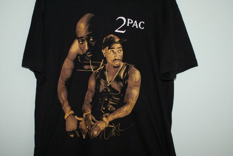 K&ouml;nnte beinhalten: Schwarzes T-Shirt mit einem goldenen und schwarzen Bild des Rappers 2Pac. Das Bild zeigt zwei verschiedene Portr&auml;ts von 2Pac, eines mit einem Bandana und eines ohne. Das Shirt hat den Text "2PAC" in Wei&szlig; auf der Vorderseite.