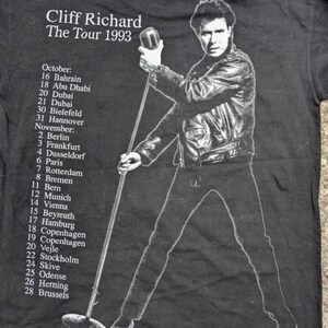 May include: Black and white photo of Cliff Richard on a black t-shirt. The t-shirt has the text "Cliff Richard The Tour 1993" and a list of tour dates in October and November 1993.