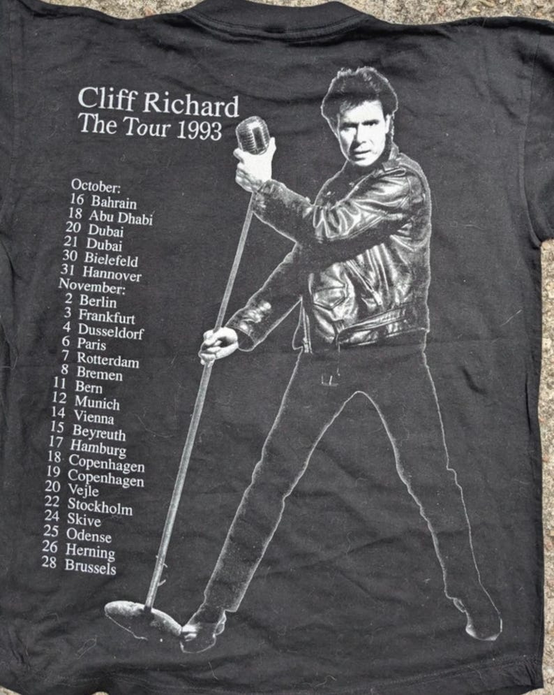 May include: Black and white photo of Cliff Richard on a black t-shirt. The t-shirt has the text "Cliff Richard The Tour 1993" and a list of tour dates in October and November 1993.