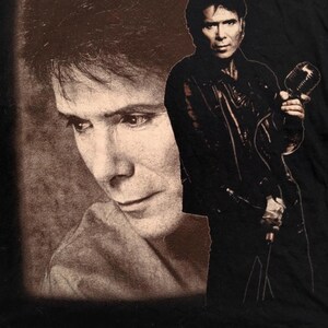 May include: A black and white photo of a man with dark hair and a leather jacket holding a microphone. The photo is printed on a black t-shirt.