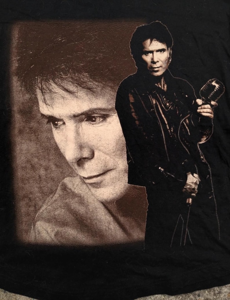 May include: A black and white photo of a man with dark hair and a leather jacket holding a microphone. The photo is printed on a black t-shirt.