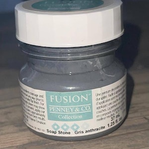 May include: A small glass jar of gray paint with a white lid. The label reads "FUSION PENNEY & CO. Collection Soap Stone Gris anthracite 1.25 fl. oz.". The jar is filled with a gray, opaque paint.