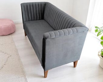 May include: A gray velvet loveseat with vertical channel tufting on the back and arms. The sofa has wooden legs and a low profile. A pink ottoman and a patterned rug are in the background.