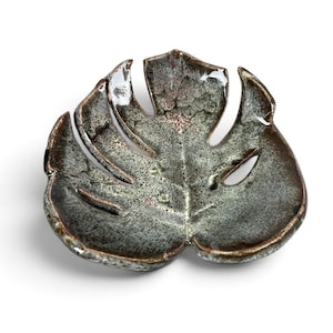 May include: A ceramic dish shaped like a monstera leaf with a glossy, mottled glaze in shades of dark green, brown, and grey. The dish features deep cutouts along the edges, mimicking the natural fenestrations of a tropical plant leaf.