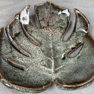 May include: A ceramic dish shaped like a monstera leaf. The dish is glazed in shades of green, brown, and white, with a speckled finish.