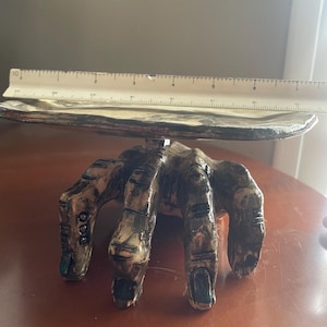 May include: A ceramic hand-shaped serving dish with a flat, round top. The hand is detailed with textured skin and dark accents on the knuckles and fingernails. A ruler is placed on the dish, showing measurements in inches.
