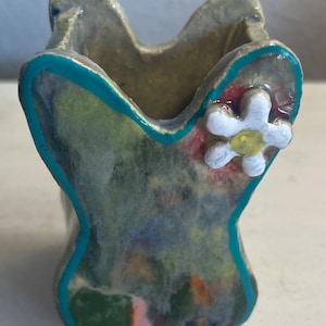 May include: A small, handmade ceramic vase with a unique shape. The vase features a turquoise outline and a colorful, abstract design. A white flower with a yellow center is attached to the side. The vase is approximately 4 inches tall.