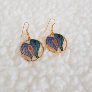 May include: Gold-coloured earrings with a floral design. Each earring has a circular frame with a purple and teal flower design. The earrings have a hook closure.