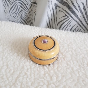 May include: A round, pale yellow trinket box with a metallic silver rim. The lid features a central purple cabochon stone surrounded by a black circular border and a ring of small, clear rhinestones set against the yellow enamel finish.