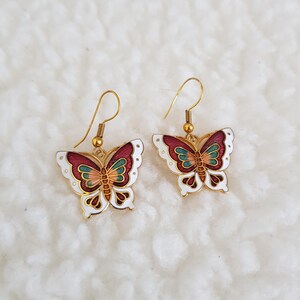 May include: A pair of butterfly earrings with gold-coloured hooks. The butterfly wings are white with red, green, and orange details. The earrings are set against a white, textured background.