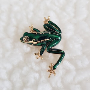 May include: A green frog brooch with gold-coloured detailing. The frog has a glossy, emerald green enamel finish and gold feet. The brooch is a decorative accessory, suitable for adding a touch of nature-inspired charm to clothing or accessories.