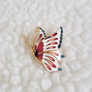 May include: A gold butterfly brooch with detailed enamel work. The wings display red, white, and blue colours, with a fine gold wire antenna. The brooch is set against a soft, white background.