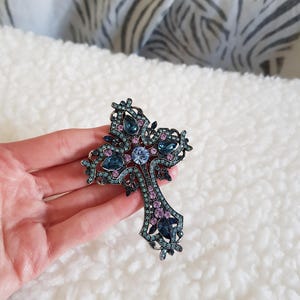 May include: An ornate cross-shaped brooch with a dark teal metal frame. The brooch is embellished with blue, purple, and clear crystals. The cross is held in a hand against a white, textured background.