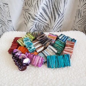 May include: A selection of colourful beaded bracelets in shades of red, orange, purple, pink, green, and blue. The bracelets are made of rectangular and oval beads. The background is a white, fluffy surface.