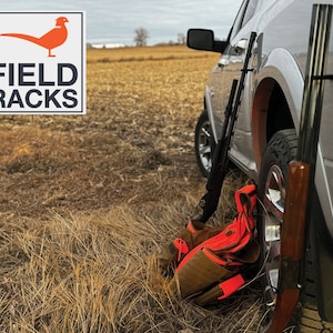 May include: Two shotguns are mounted on the back of a silver pickup truck using a Field Racks gun rack. A brown hunting vest with orange accents is lying on the ground next to the truck.