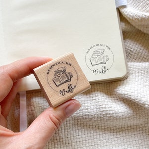 Ein Holzstempel mit dem Motiv "Aus dem Regal von" und dem Namen als Personalisierung. In der Mitte des Stempelmotivs ist ein Stapel Bücher zu sehen.