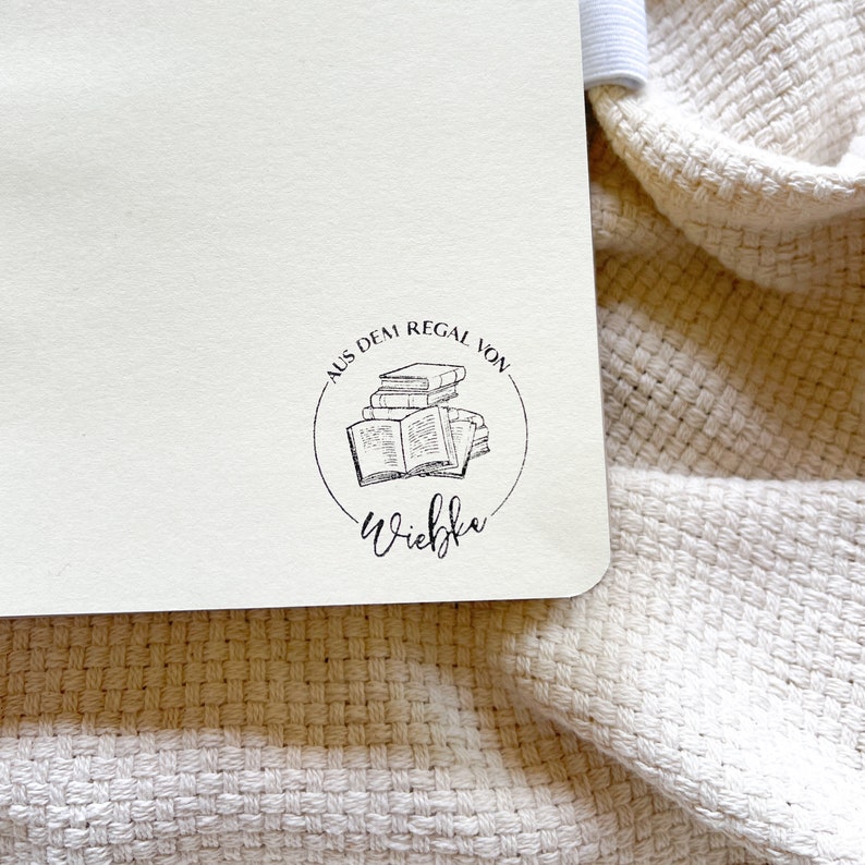 Ein Holzstempel mit dem Motiv "Aus dem Regal von" und dem Namen als Personalisierung. In der Mitte des Stempelmotivs ist ein Stapel Bücher zu sehen.