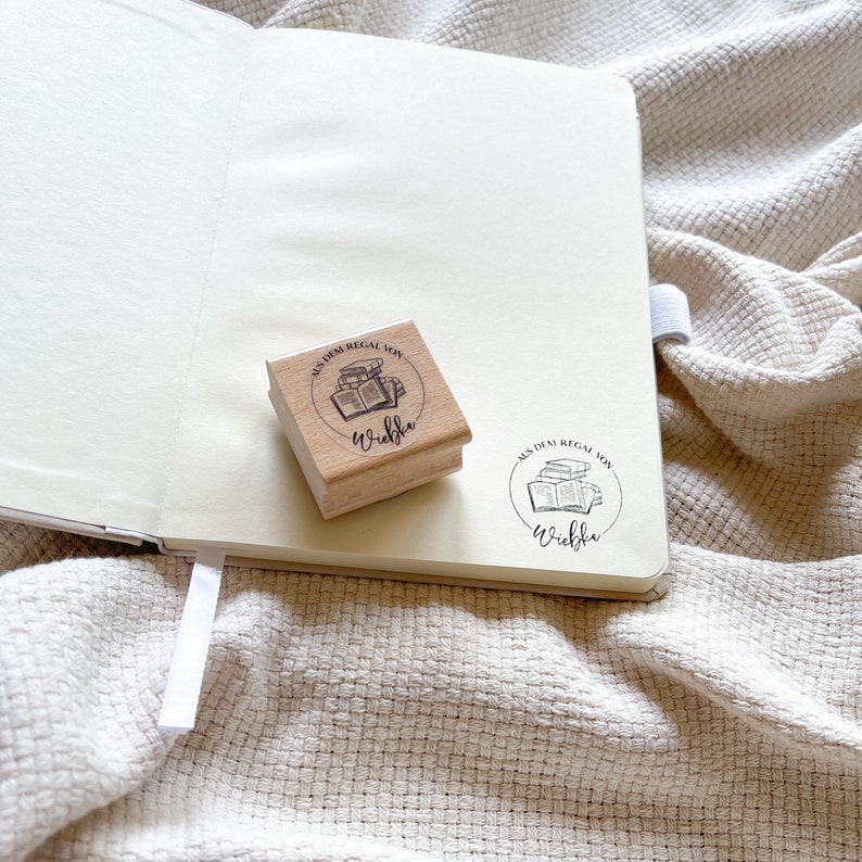 Ein Holzstempel mit dem Motiv "Aus dem Regal von" und dem Namen als Personalisierung. In der Mitte des Stempelmotivs ist ein Stapel Bücher zu sehen.