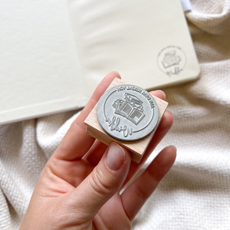 Ein Holzstempel mit dem Motiv "Aus dem Regal von" und dem Namen als Personalisierung. In der Mitte des Stempelmotivs ist ein Stapel Bücher zu sehen.