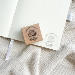 Ein Holzstempel mit dem Motiv "from the shelf of" und dem Namen als Personalisierung. In der Mitte des Stempelmotivs ist ein Stapel Bücher zu sehen.