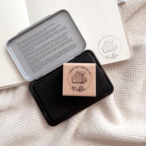 Ein Holzstempel mit dem Motiv "from the shelf of" und dem Namen als Personalisierung. In der Mitte des Stempelmotivs ist ein Stapel Bücher zu sehen. Darunter liegt ein schwarzes Stempelkissen.