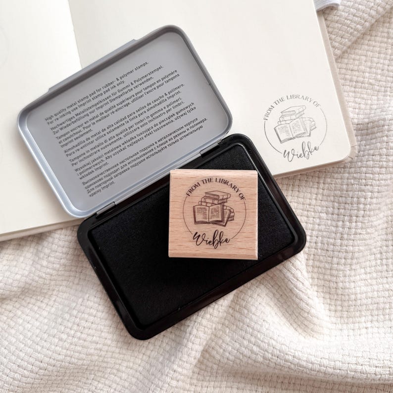 Ein Holzstempel mit dem Motiv "from the shelf of" und dem Namen als Personalisierung. In der Mitte des Stempelmotivs ist ein Stapel Bücher zu sehen. Darunter liegt ein schwarzes Stempelkissen.