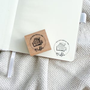 Ein Holzstempel mit dem Motiv "Aus dem Regal von" und dem Namen als Personalisierung. In der Mitte des Stempelmotivs ist ein Stapel Bücher zu sehen.