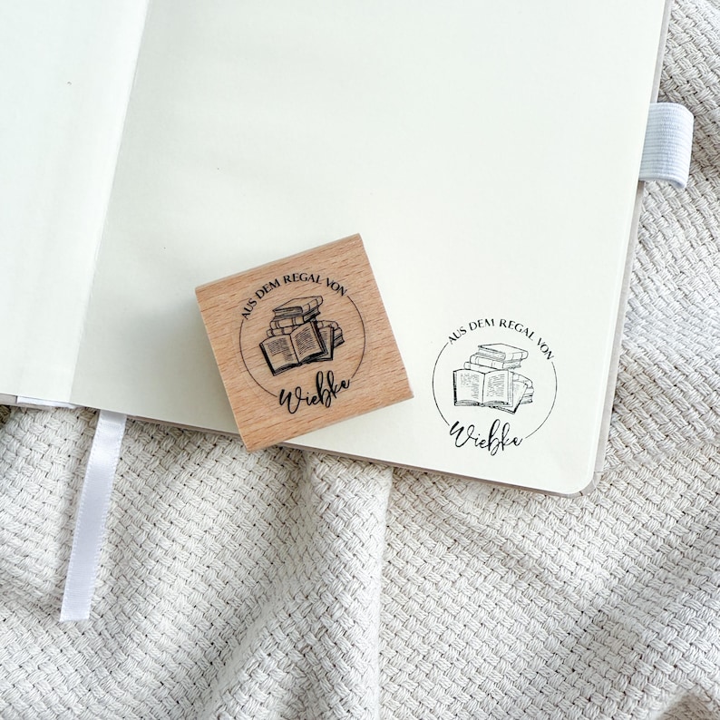 Ein Holzstempel mit dem Motiv "Aus dem Regal von" und dem Namen als Personalisierung. In der Mitte des Stempelmotivs ist ein Stapel Bücher zu sehen.