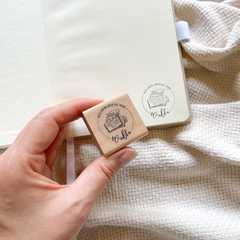 Ein Holzstempel mit dem Motiv "Aus dem Regal von" und dem Namen als Personalisierung. In der Mitte des Stempelmotivs ist ein Stapel Bücher zu sehen.