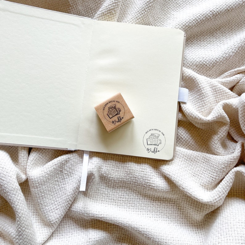 Ein Holzstempel mit dem Motiv "Aus dem Regal von" und dem Namen als Personalisierung. In der Mitte des Stempelmotivs ist ein Stapel Bücher zu sehen.
