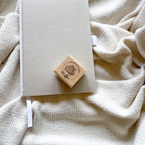 Ein Holzstempel mit dem Motiv "Aus dem Regal von" und dem Namen als Personalisierung. In der Mitte des Stempelmotivs ist ein Stapel Bücher zu sehen.