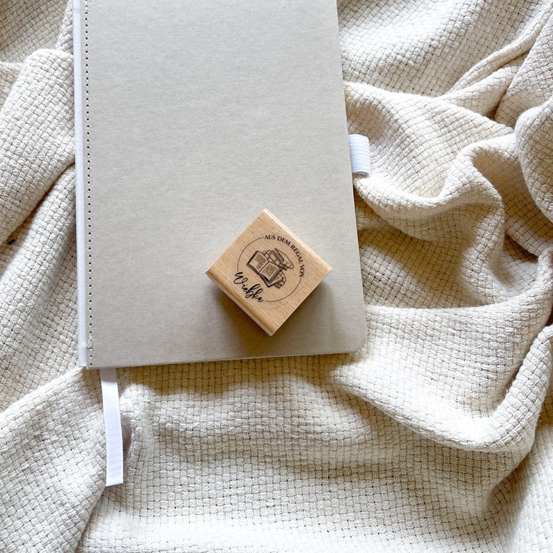 Ein Holzstempel mit dem Motiv "Aus dem Regal von" und dem Namen als Personalisierung. In der Mitte des Stempelmotivs ist ein Stapel Bücher zu sehen.