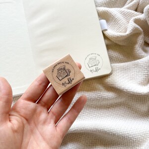 Ein Holzstempel mit dem Motiv "Aus dem Regal von" und dem Namen als Personalisierung. In der Mitte des Stempelmotivs ist ein Stapel Bücher zu sehen.