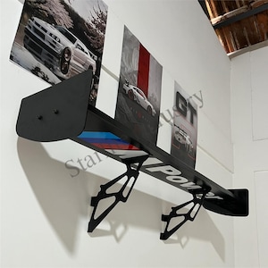 May include: Black wall shelf in the shape of a car spoiler, mounted on a white wall. The shelf has the text "M Power" in white. The shelf is decorated with a blue, red, and white stripe. Three car posters are mounted above the shelf.