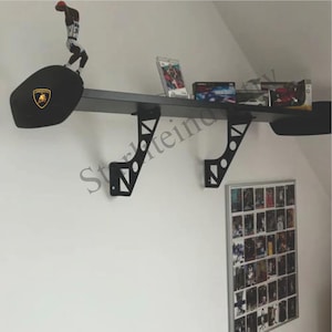 May include: Black wall shelf designed to resemble a car spoiler, with a Lamborghini logo. The shelf is mounted on a white wall and holds various items, including a figurine, video games, and model cars.