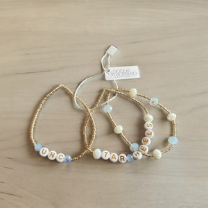 May include: Three gold beaded bracelets with white letter beads spelling out the words "UNC", "TAR", and "HEELS". The bracelets are on a light brown wooden surface.
