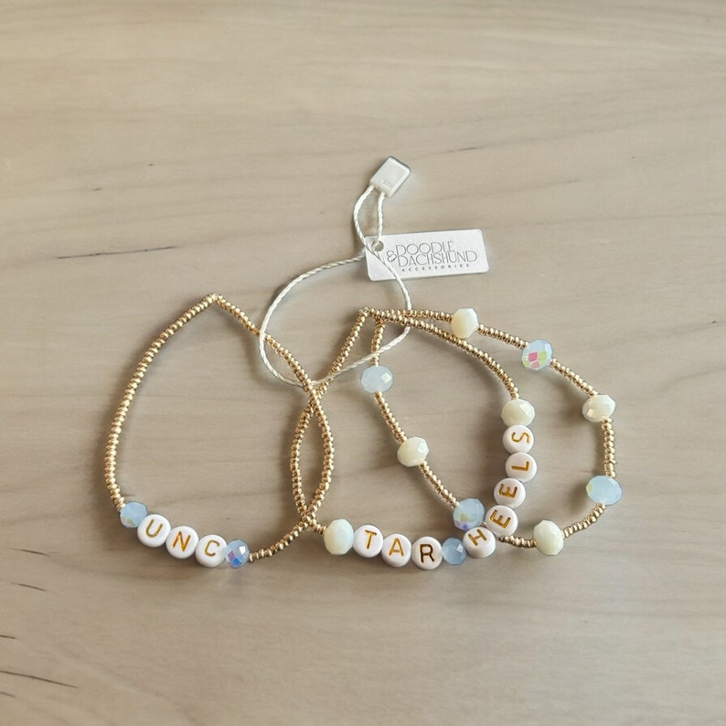 May include: Three gold beaded bracelets with white letter beads spelling out the words "UNC", "TAR", and "HEELS". The bracelets are on a light brown wooden surface.