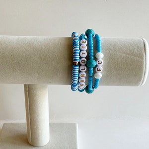 May include: A set of five bracelets with blue and white beads. The bracelets are stacked on a white display stand. One bracelet has the word "CHARLOTTE" spelled out in gold letters on white beads. Another bracelet has the letters "FC" spelled out in gold letters on white beads.
